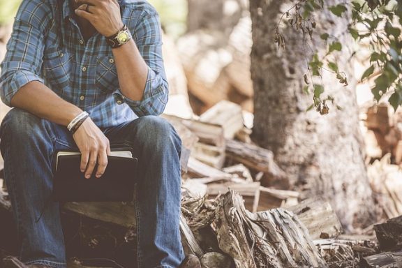 Person sitzt auf einem Holzscheit und hält ein Buch, umgeben von Holzstapeln.