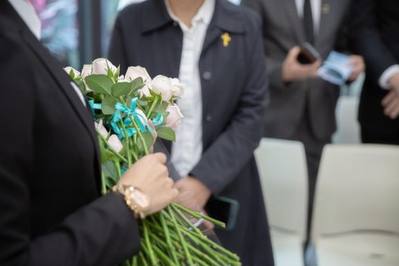 Eine Person hält einen Strauß weißer Rosen in der Hand, im Hintergrund eine Gruppe von Menschen.