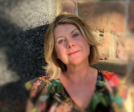 Eine Frau mit blonden, lockigen Haaren und einem bunten Top lächelt sanft im Freien.