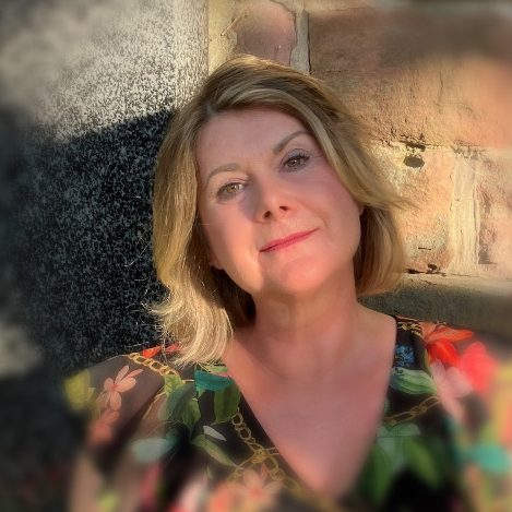 Frau mit blonden Haaren, in buntem Oberteil, blickt freundlich in die Kamera.