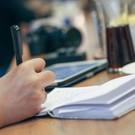 Jemand schreibt in ein Notizbuch, während Getränke und ein Tablet auf dem Tisch stehen.